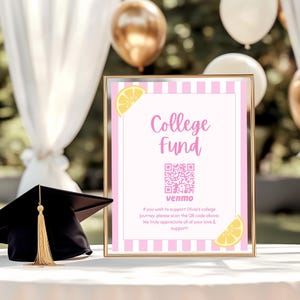 May include: A framed sign with the words "College Fund" in pink script, a QR code, and the Venmo logo. The sign is decorated with pink stripes and lemon illustrations. A black graduation cap with a gold tassel sits on a table.