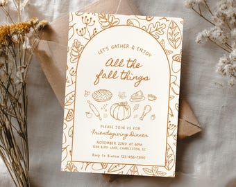 Puede incluir: Una invitación de Friendsgiving de color crema con texto e ilustraciones marrones con temática otoñal. El texto dice "Let's Gather & Enjoy All the Fall Things" e incluye la fecha, la hora y el lugar. Hay un sobre marrón detrás de la invitación.