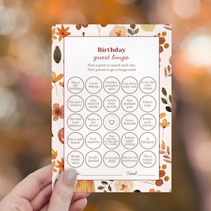 May include: A fall-themed birthday guest bingo card with floral accents. The card features various prompts like "Loves baking" and "Has a pet hamster." The text "Birthday guest bingo" is at the top.