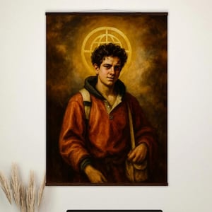May include: A wall hanging featuring a portrait of a person in an orange jacket with a halo. The artwork is displayed above a wooden desk with a computer, accessories, and a gray blanket draped over a chair.