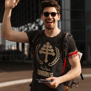 Peut inclure: Un homme portant un t-shirt noir avec un motif doré d'une croix et le texte "Maronite" en arabe. Il porte des lunettes de soleil et sourit, saluant de la main droite.