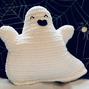 May include: A handmade white crochet ghost with a smiling face and outstretched arms. The ghost has a black mouth and eyes. The background is a dark blue pillow with a white spiderweb and spider design. A Halloween-themed item.