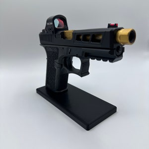 May include: A black handgun with a gold barrel and a red dot sight. The gun is on a black stand.