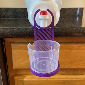 Può includere: Dispenser in plastica viola con un pulsante rosso e una tazza trasparente attaccata. Il dispenser è montato su una brocca bianca.