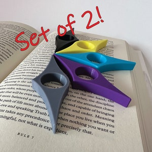 May include: Set of two colorful bookmark holders in the shape of a star. The bookmarks are made of plastic and are in the colors purple, blue, yellow, and gray.