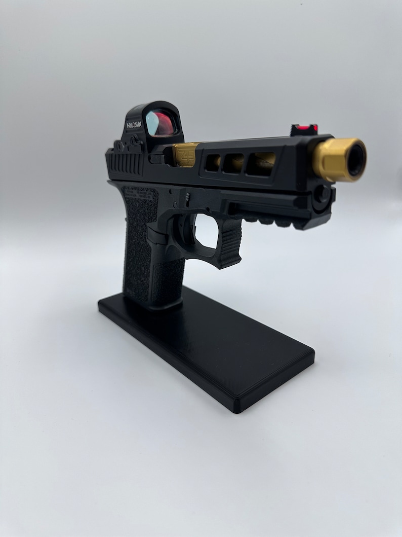May include: A black handgun with a gold barrel and a red dot sight. The gun is on a black stand.
