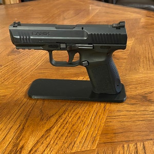 May include: A black handgun with a silver slide and a red safety lever is displayed on a black stand. The gun is on a wooden table.