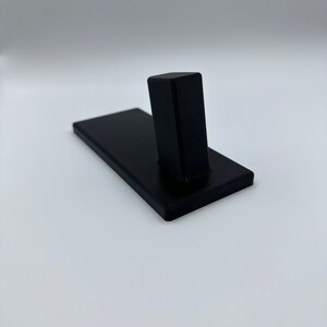 May include: A black plastic stand with a rectangular base and a vertical post. The stand is designed to hold a small object, such as a phone or a tablet.