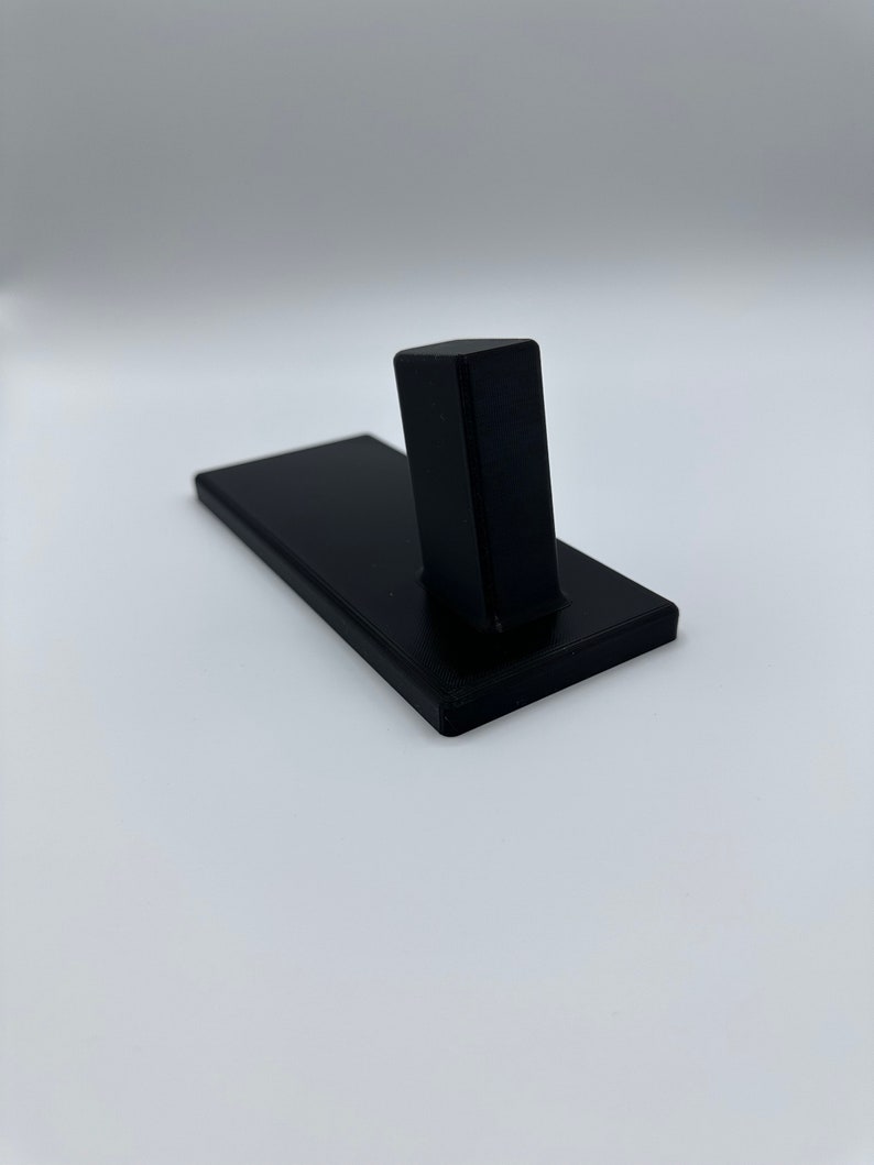 May include: A black plastic stand with a rectangular base and a vertical post. The stand is designed to hold a small object, such as a phone or a tablet.