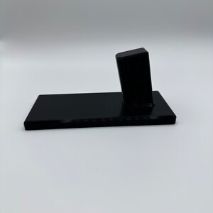 May include: A black plastic phone stand with a rectangular base and a raised rectangular platform. The stand is designed to hold a phone upright.