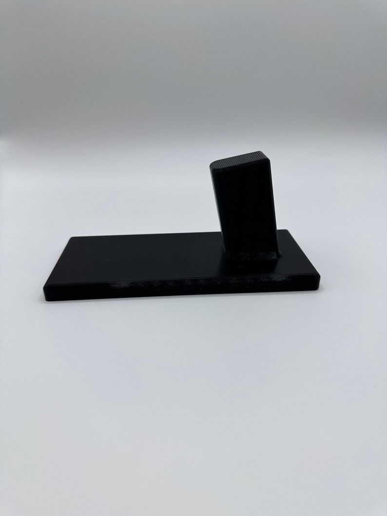 May include: A black plastic phone stand with a rectangular base and a raised rectangular platform. The stand is designed to hold a phone upright.