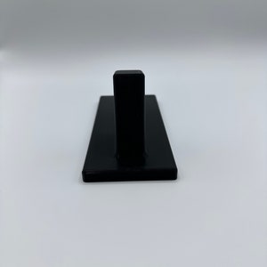 May include: A black plastic wall hook with a single hook on the top. The hook is mounted on a rectangular base.