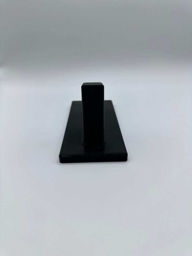 May include: A black plastic wall hook with a single hook on the top. The hook is mounted on a rectangular base.