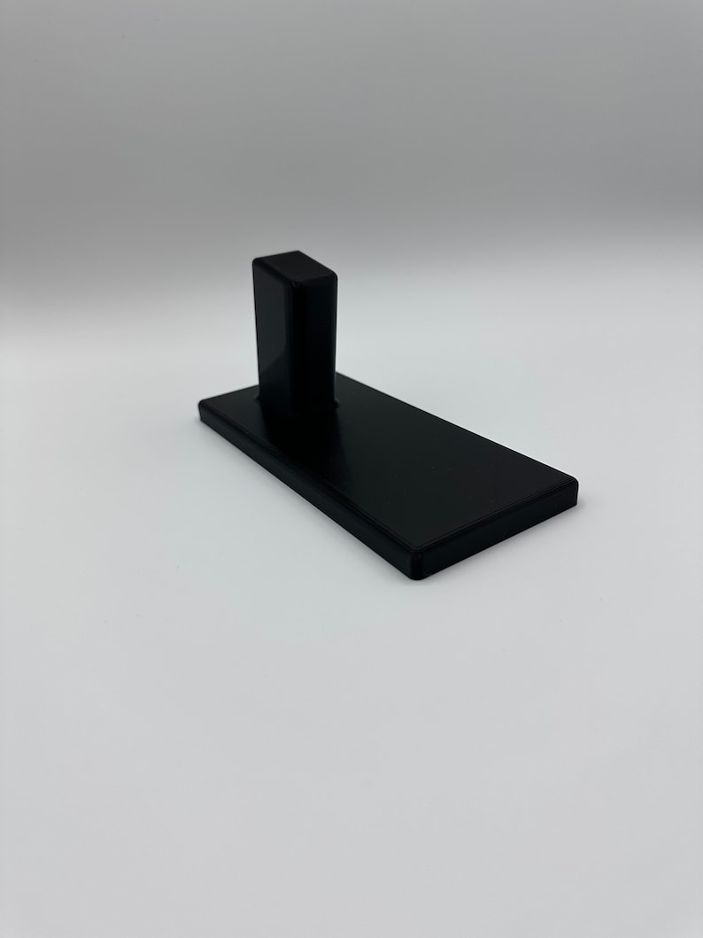 May include: A black plastic stand with a raised platform. The stand is designed to hold a small object, such as a phone or a tablet.