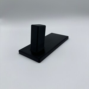 May include: A black wall-mounted hook with a rectangular base and a single vertical hook.