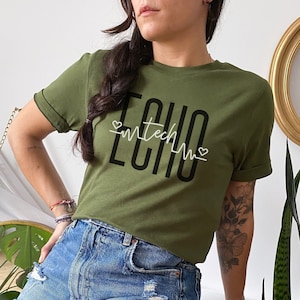 Puede incluir: Una mujer con una camiseta verde con el texto "ECHO" y un gráfico de latido del corazón blanco. Lleva pantalones cortos de mezclilla y tiene un tatuaje en el brazo.