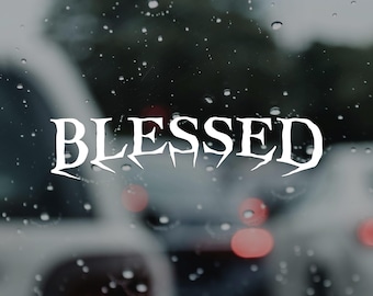 May include: White text that reads "BLESSED" in a stylized font. The image is taken through a window with raindrops, with blurred background of cars and traffic lights.