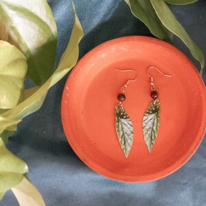 May include: A pair of dangle earrings with green leaf-shaped charms and brown beads. The earrings are on a terracotta-colored plate.