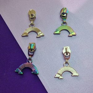 May include: Four iridescent rainbow zipper pulls with a metal slider and a rainbow-shaped charm. The zipper pulls are on a white and purple background.