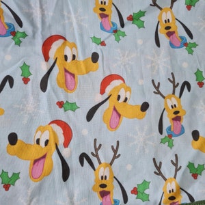 May include: Pale blue fabric featuring a repeating pattern of Pluto's face wearing a Santa hat and reindeer antlers. The design includes snowflakes and holly leaves, creating a festive holiday theme.