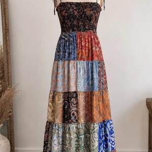 Könnte beinhalten: Ein ärmelloses Maxikleid mit gesmoktem Oberteil und Trägern zum Binden. Der Rock besteht aus mehreren horizontalen Stoffbahnen, die jeweils unterschiedliche Paisley- und Blumenmuster in Rot-, Blau- und Brauntönen aufweisen.
