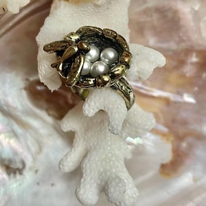 Könnte beinhalten: Ein Ring mit einem strukturierten Vogelnest-Design aus antik goldfarbenem Metall. Das Nest enthält drei kleine, runde, weiße Kunstperlen und ist mit einem kleinen Metallvogel am Rand verziert. Die Ringschiene hat eine passende strukturierte Oberfläche.