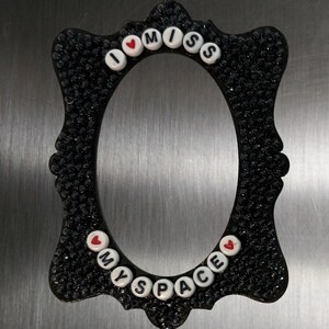 May include: An oval-shaped black picture frame with a textured, sparkling surface. The frame is decorated with white plastic alphabet beads spelling out the phrase I MISS MY SPACE, interspersed with small red heart symbols on the top and bottom.