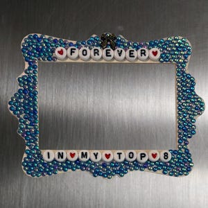 May include: A rectangular picture frame decorated with iridescent blue beads and a small black bow at the top. The frame features white letter beads spelling "FOREVER" at the top and "IN MY TOP 8" at the bottom, interspersed with small red heart beads.