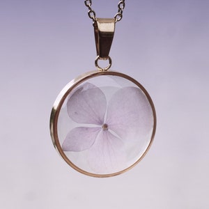 Peut inclure: Un collier pendentif doré avec un médaillon rond en verre transparent. À l'intérieur du médaillon, une délicate fleur violet clair. La chaîne du collier est en métal fin argenté. Le fond est un dégradé doux de violet et de blanc.