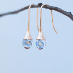 Peut inclure: Boucles d'oreilles pendantes dorées. Chaque boucle d'oreille présente un globe de verre transparent en forme de goutte d'eau contenant une petite fleur bleue et jaune. Les boucles d'oreilles sont suspendues à un crochet incurvé.
