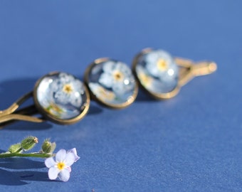May include: Three gold cufflinks with forget-me-not flowers pressed inside clear resin. The cufflinks are on a blue surface with a single forget-me-not flower.