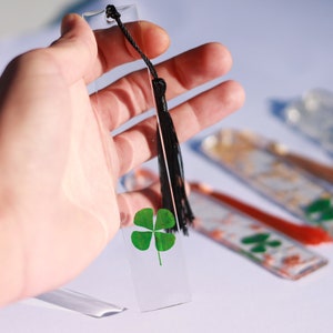 Peut inclure: Un marque-page en verre transparent avec un trèfle à quatre feuilles et un pompon noir. Le marque-page est tenu dans une main.