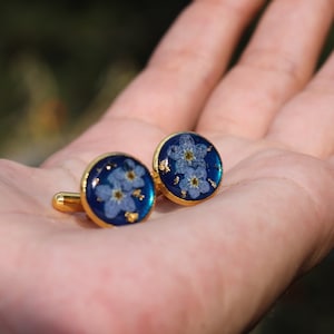 以下が含まれることがあります： 青いレジンと押し花されたワスレナグサの花が付いた、金色のカフスボタンのペアです。