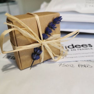 May include: A small brown gift box tied with a natural raffia ribbon and decorated with dried lavender sprigs. The box is sitting on a white surface with a business card that says "Idees de France" and "75012 Paris".