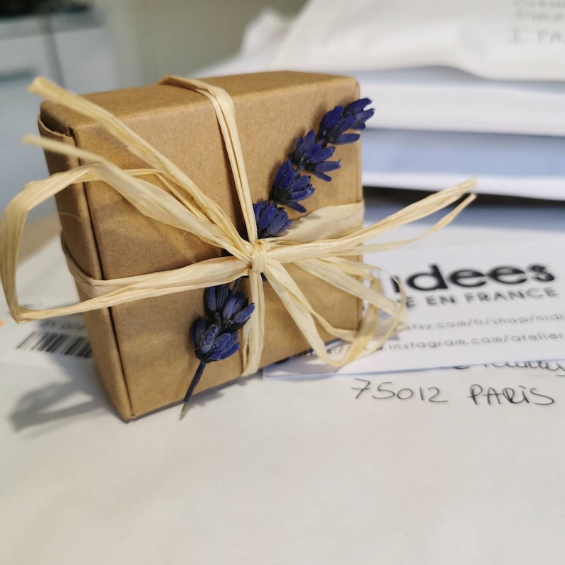 May include: A small brown gift box tied with a natural raffia ribbon and decorated with dried lavender sprigs. The box is sitting on a white surface with a business card that says "Idees de France" and "75012 Paris".
