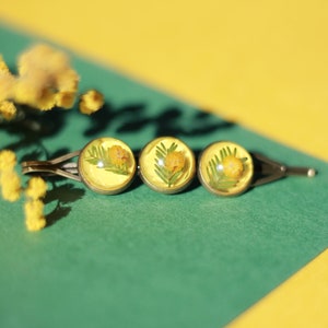 May include: A hair barrette with three circular, clear resin settings, each containing a yellow flower and green leaves. The barrette is set against a green and yellow background, with a sprig of yellow flowers.