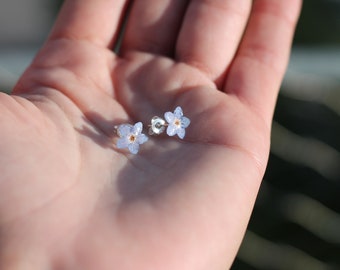 May include: A pair of silver stud earrings featuring delicate forget-me-not flowers pressed in resin. The flowers are a light blue color with a yellow center.