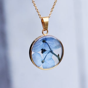 Puede incluir: Un collar con colgante dorado con un dije circular de resina transparente. El dije contiene flores azules. El collar tiene una fina cadena dorada y un cierre sencillo. El fondo es un suave desenfoque blanco y azul.