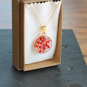 Peut inclure: Un collier en or avec un pendentif rond présentant des fleurs pressées rouges enchâssées dans de la résine transparente. Le collier est présenté dans une boîte brune avec une doublure blanche.