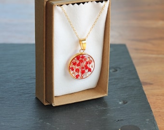 May include: A gold necklace with a round pendant featuring red pressed flowers encased in clear resin. The necklace is displayed in a brown box with white lining.