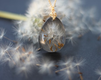 May include: A gold chain necklace with a teardrop-shaped pendant filled with a delicate dandelion seed head. The pendant is surrounded by several dandelion seeds.