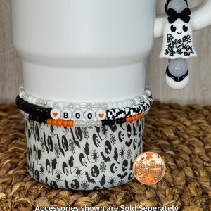May include: A white tumbler with a black and white Halloween-themed design. The tumbler has a beaded bracelet that spells out "BOO" in orange and white beads. A ghost-shaped charm is attached to the handle. The accessories shown are sold separately.