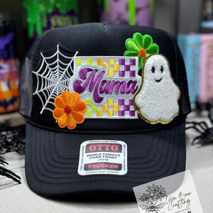 May include: Black trucker hat with a variety of colorful patches, including a ghost, a spiderweb, a flower, and a checkered patch with the word "Mama" in purple. The hat has a black mesh back and a label that reads "OTTO World Famous Foam Front OSFM Trucker".