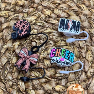 May include: Four colorful shoe charms with black and white strings. The charms include a football helmet with a leopard print, a pink bow with white stitching, a rainbow colored "CHEER mama" charm, and a black and white "MAMA" charm with a lightning bolt.