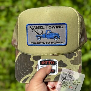 May include: A green and brown camouflage trucker hat with a patch that says "CAMEL TOWING" and "WE'LL GET YOU OUT OF A PINCH". The patch features a blue tow truck with a white background.