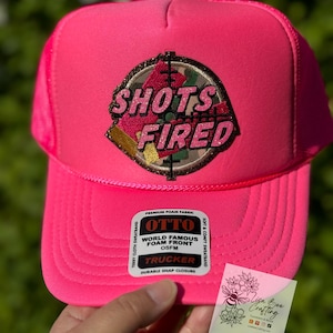 May include: Pink trucker hat with a black and green camouflage patch that says "Shots Fired" in pink glitter letters. The hat has a black mesh back and a white label that says "Otto World Famous Foam Front OSFM Trucker Durable Snap Closure".