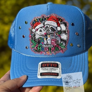 May include: A light blue trucker hat with a mesh back and a curved brim. The front features an embroidered patch with a raccoon and opossum wearing Santa hats and the text "Trashy Christmas". The hat is decorated with colorful rhinestones.