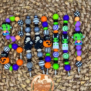 May include: A collection of Halloween-themed beaded pens in various colors. The pens feature decorative beads with designs including ghosts, pumpkins, Frankenstein's monster, and checkered patterns. The pens are arranged on a woven surface.