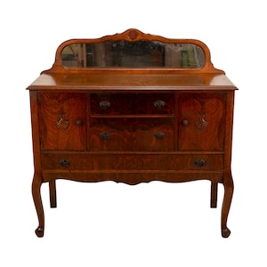 May include: A vintage wooden sideboard with a mirror. The dark brown cabinet has two doors, three drawers, and a decorative mirror. The cabinet has curved legs and ornate hardware. The wood grain is visible throughout the piece.