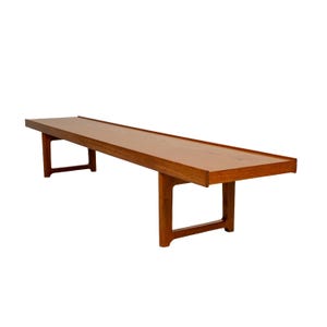May include: A long, rectangular wooden coffee table with a warm brown finish. The table has a raised edge and is supported by four sturdy, angled legs. The design is mid-century modern, with clean lines and a minimalist aesthetic.